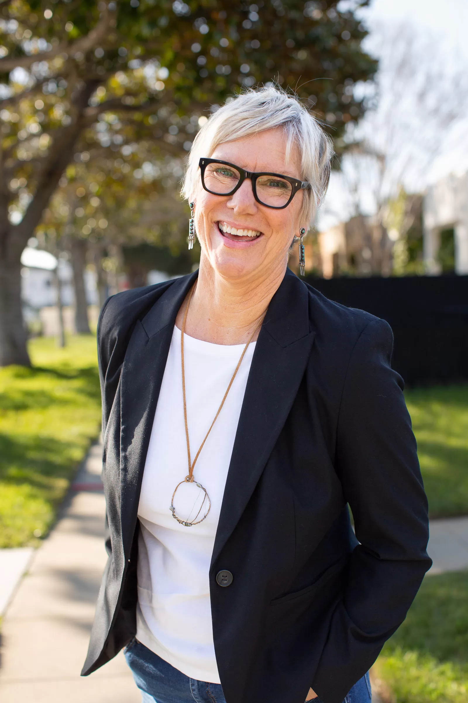 A woman with short gray hair and glasses is smiling while standing on a sidewalk. She is wearing a black blazer over a white shirt and a necklace with a large round pendant. Trees and grass are in the background under a sunny sky.