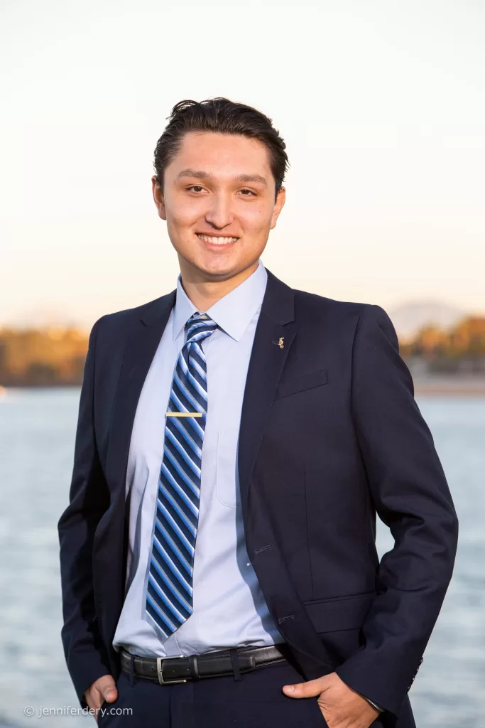 president of the pacific beach town council stands outdoors near a body of water, smiling at the camera. He is wearing a dark blue suit with a light blue dress shirt and a blue striped tie. His hands are in his pockets, and the background is slightly blurred, showing some trees and distant land.