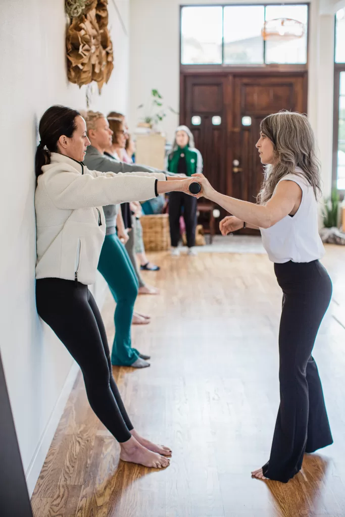 A group of people in a spacious, well-lit room practice an exercise against a wall. One individual helps another by guiding their outstretched arm, ensuring proper form. Others wait their turn along the wall. The atmosphere appears calm and focused.