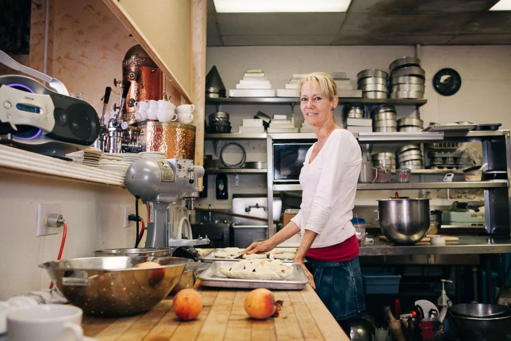 Michelle Coulon of dessertier.com stands in a commercial kitchen, smiling and preparing pastries on a wooden countertop. Various kitchen equipment, including mixing bowls and baking pans, can be seen in the background. Two peaches are resting on the countertop in the foreground.