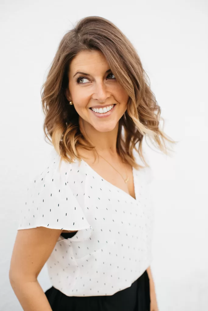 A woman with shoulder-length wavy hair smiles while looking to her left. She is wearing a white blouse with small black dots and a black skirt. The background is plain white.