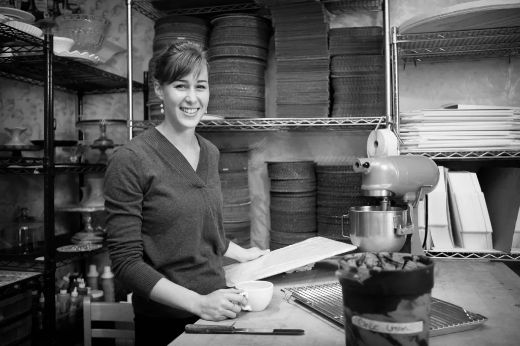 A woman stands in a bakery or kitchen, smiling while holding a cup in one hand and a baking tray in the other. Shelves filled with baskets, plates, and various kitchen items surround her. A stand mixer and a container of ingredients are on the counter. The image is in black and white.