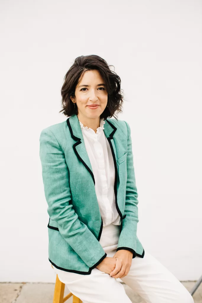 A woman with shoulder-length dark hair sits on a wooden stool against a white background. She is wearing a light green blazer with black trim over a white blouse and white pants. She has a slight smile and her hands rested on her lap.