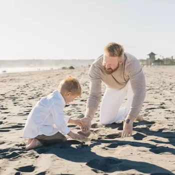 photo of a father and son playing in the sand on Coronado beach during a San Diego Family Portrait Session. both are wearing tan pants with white button down shirts and have redish hair.
