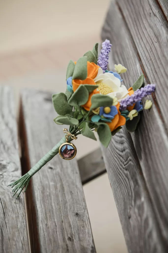 diy handmade felt flower bridal bouquet in green, blue, purple, white, and orange. locket tied to bouquet contains a photo of cherished family cat