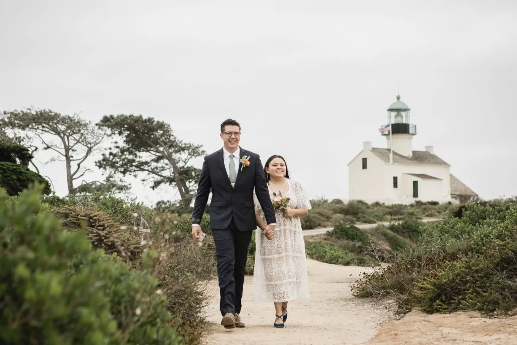 elopement photo of a bride and groom walking cliffside in san diego at cabrillo monument park with the the lighthouse in the background
