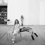 A black and white photo by a family photographer captures three young girls. Two girls sit on a bench, one with a relaxed pose and the other lying down. A third girl stands at a distance behind the bench, smiling. There's a plant stand and a storefront in the background.