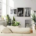 A bright living room with large windows allowing natural light. The space features a beige sectional sofa, several green plants, and three black-and-white photos on the wall, including a wedding album display. There is also a small wooden coffee table and a modern black chair.