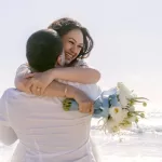 A smiling woman in a white dress is being lifted and hugged by a person in a white shirt on a sunny San Diego beach. She holds a bouquet of white flowers, and they both appear joyful with the ocean and clear sky, capturing the essence of wedding photography.