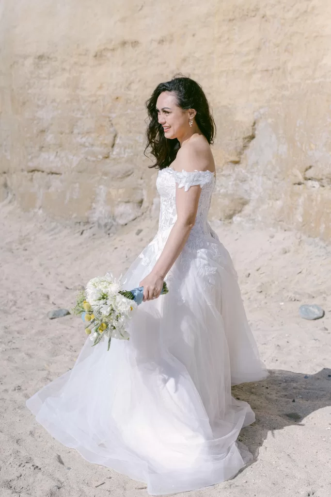 bride in a white wedding dress shown in direct full-sun lighting. located at the beach in southern California. with a light tan cliff as the background