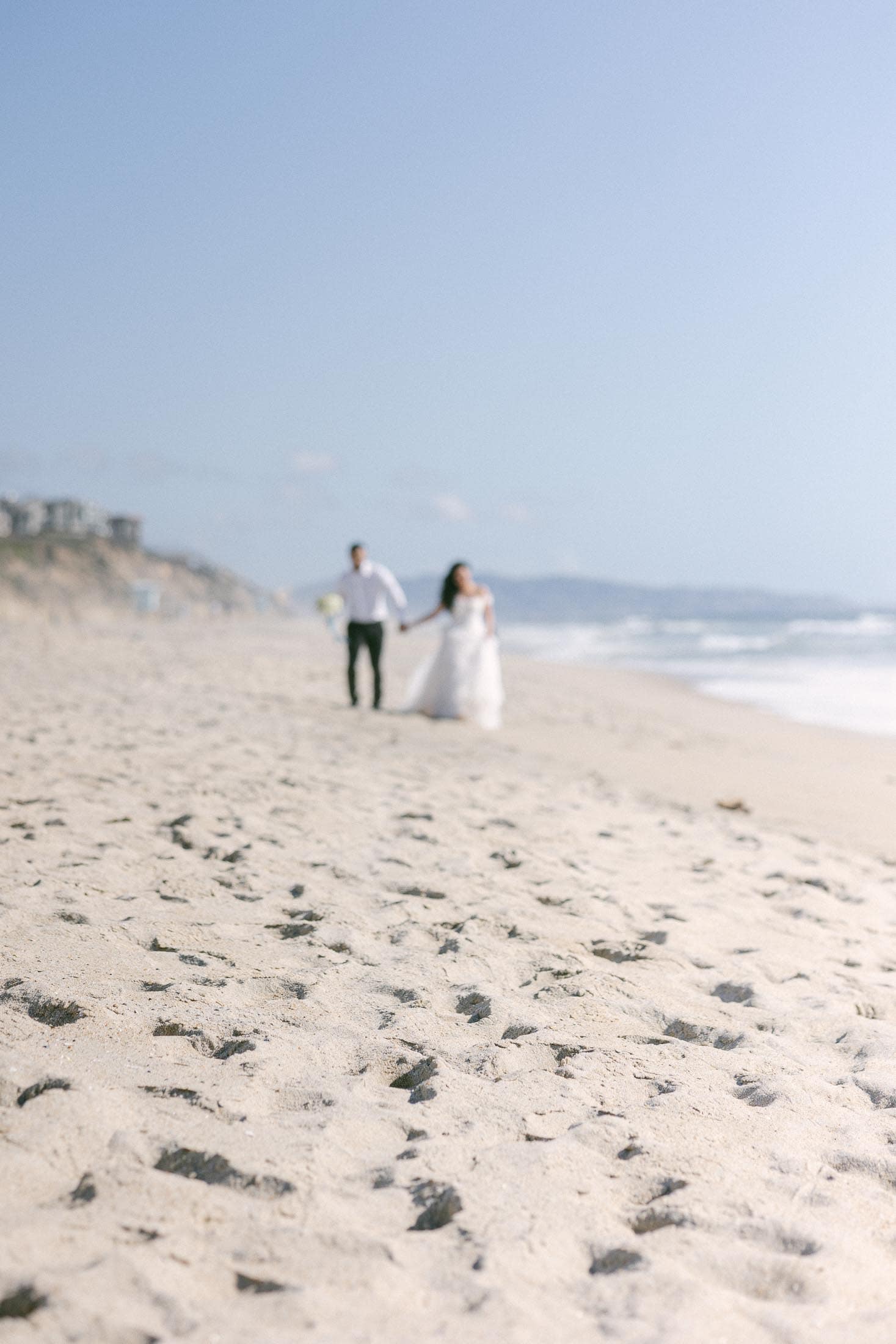 A bride and groom walk hand in hand on a sandy beach with the ocean and hills in the background, capturing the quintessential essence of wedding photography. The image is softly focused, creating a dreamy, ethereal effect. The bride wears a white dress, and the groom is dressed in a white shirt and dark pants.