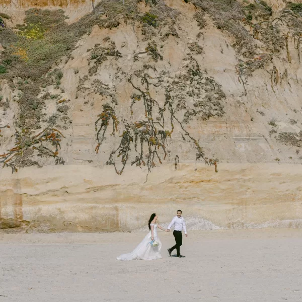 A couple, dressed in wedding attire, walks hand in hand on a sandy beach. The bride wears a flowing white gown, holding a bouquet, and the groom wears a white shirt and black pants. Behind them, a large cliff with sparse vegetation rises against a blue sky—perfect for stunning wedding photography.