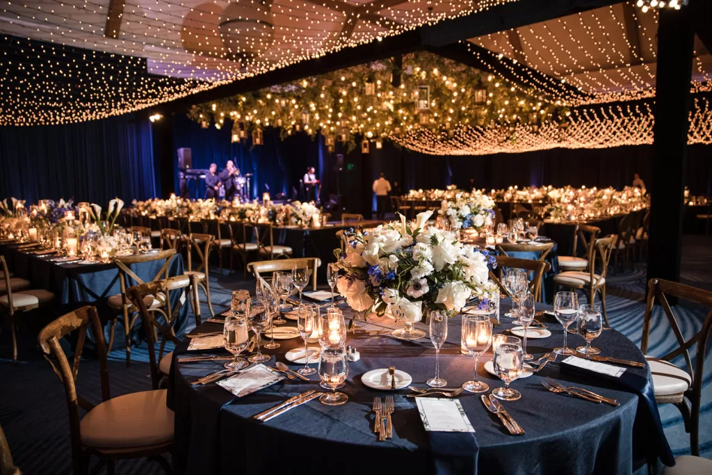 The Southpointe Ballroom is a lavishly decorated event hall at the hotel del coronado, featuring round tables set for dining with elegant floral centerpieces, all illuminated by numerous small, twinkling lights strung across the ceiling, creating a warm and enchanting atmosphere.