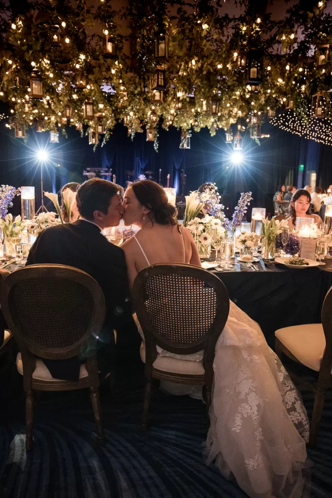 couple kissing at the head table. white floral arrangements, fairy lights, candles, and navy draped ballroom of southpointe ballroom at the hotel del coronado, CA