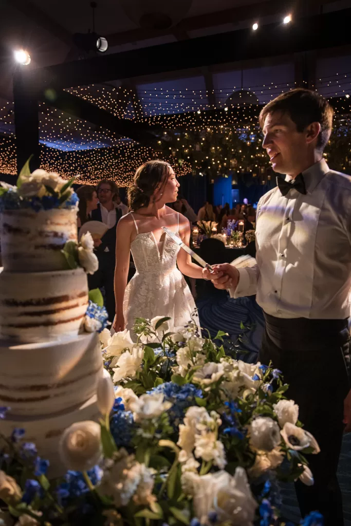 Couple waiting to cut their cake. white floral arrangements, rough-frosted wedding cake, candles, and navy draped ballroom of southpointe ballroom at the hotel del coronado, CA