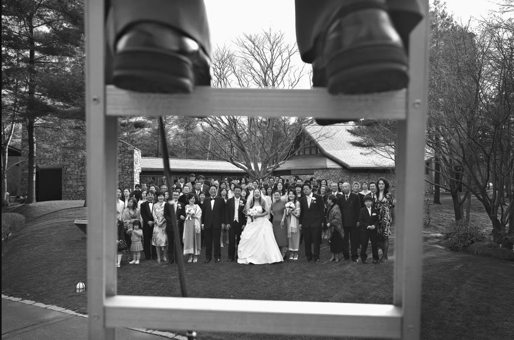 Black and white photo of a large wedding group posed outside. The bride and groom stand in the center, surrounded by their guests. The photo, showcasing classic wedding photography in japan, is taken from behind a ladder, with the photographer's shoes visible on the ladder steps. Trees and buildings fill the background.