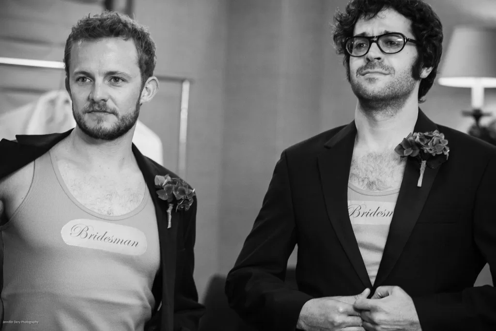 Two men stand side by side dressed in suits with open jackets, revealing tank tops that say “Bridesman”. Each has a boutonnière on their jacket lapel. One man has short hair and a beard, the other has curly hair and glasses. The setting appears formal.