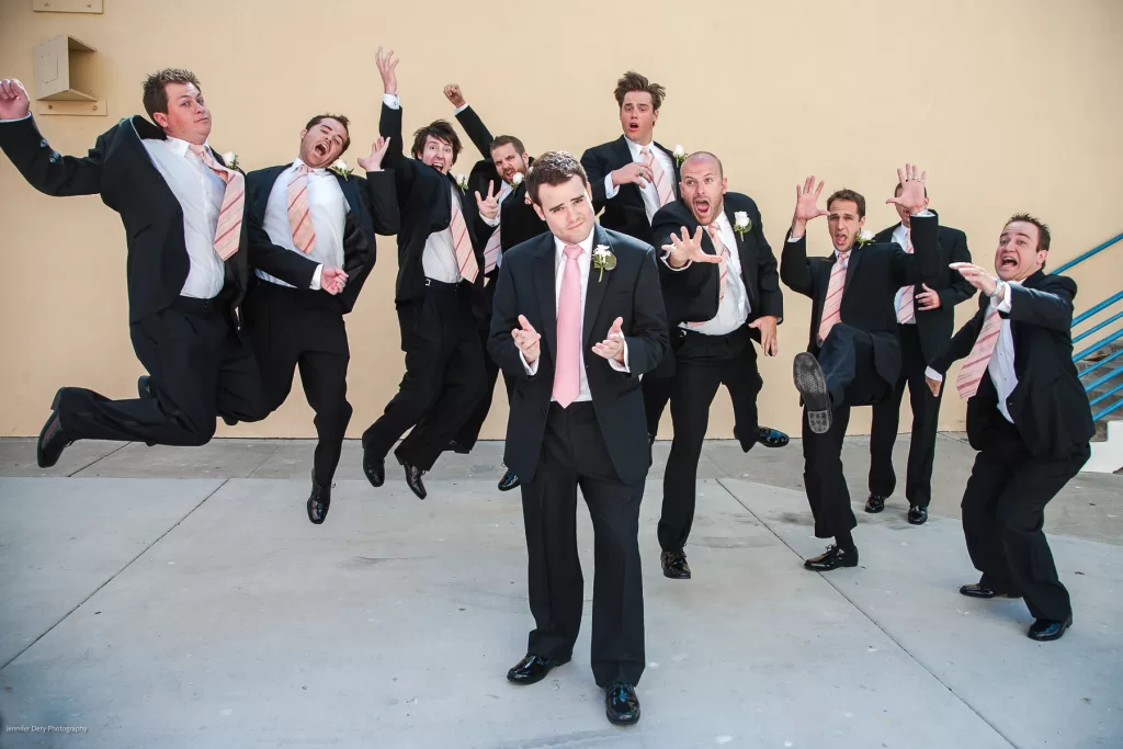 A man in a suit, standing front and center, gazes at the camera with a serious expression. Behind him, eight men in suits jump and pose energetically, creating a lively, fun atmosphere. The background is a plain, light-colored wall with a blue handrail to the right.