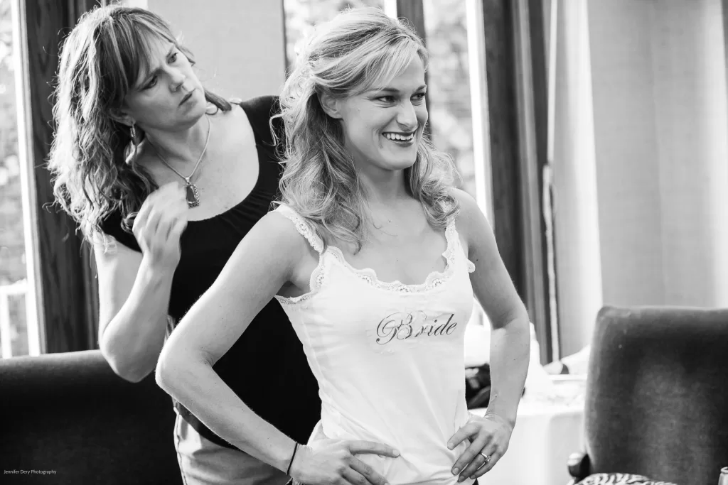 A smiling bride, wearing a tank top with "Bride" written on it, stands with her hands on her hips while a hairstylist works on her hair. They are inside a brightly lit room with large windows.