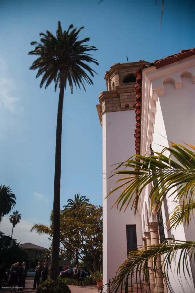 A tall palm tree stands next to a white Spanish-style building with a red-tiled roof and arched windows. The sky is clear and blue. People are gathered near the entrance of the building.