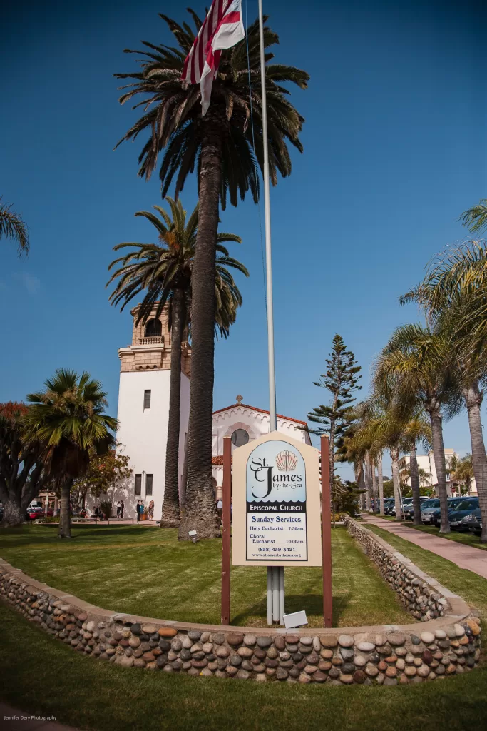 A tall palm tree and an American flag stand in front of St. James Episcopal Church, a white building with a tower, on a bright sunny day. The wooden sign in the foreground lists Sunday service times and contact information, surrounded by a stone-bordered lawn and pathway.