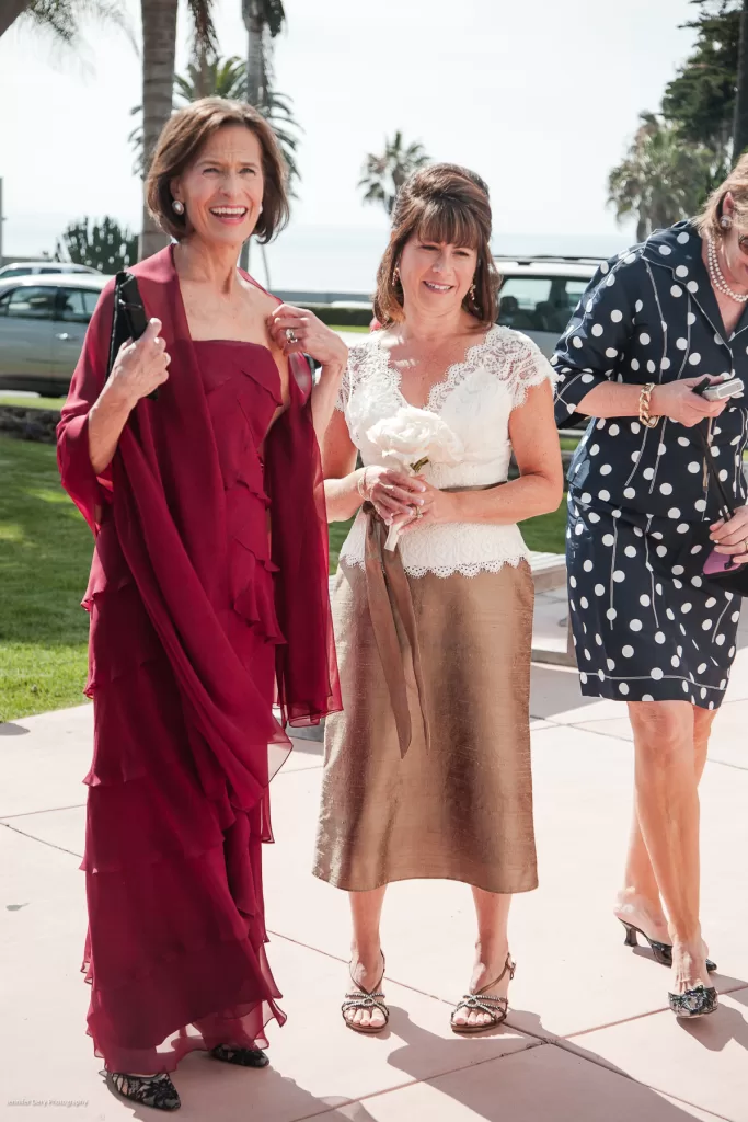 Three women are smiling and walking outdoors. The first wears a red strapless dress with a matching wrap, the second wears a white lace top and gold skirt while holding a bouquet, and the third wears a navy blue dress with white polka dots. Palm trees are in the background.