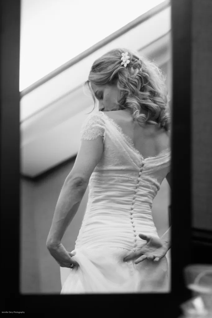 A bride, seen from behind, adjusts her white lace wedding dress in front of a mirror. Her blonde hair is styled in loose curls with a small flower accessory. The image, in black and white, captures the elegance and delicate details of her gown.