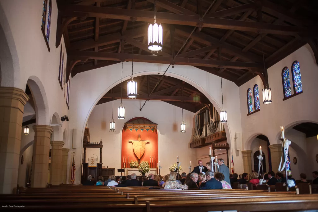 A spacious and well-lit church interior with vaulted wooden ceilings, stained glass windows, and a large archway leading to the altar. The congregation is seated in wooden pews, and some people are gathered near the entrance. The altar is adorned with religious symbols.