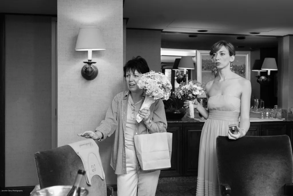 A woman in casual attire, holding a bag, stands next to a woman in a strapless dress, holding a bouquet. Both are indoors near upholstered chairs and lamps on the wall behind them. The scene appears candid and they seem to be preparing for a formal event.