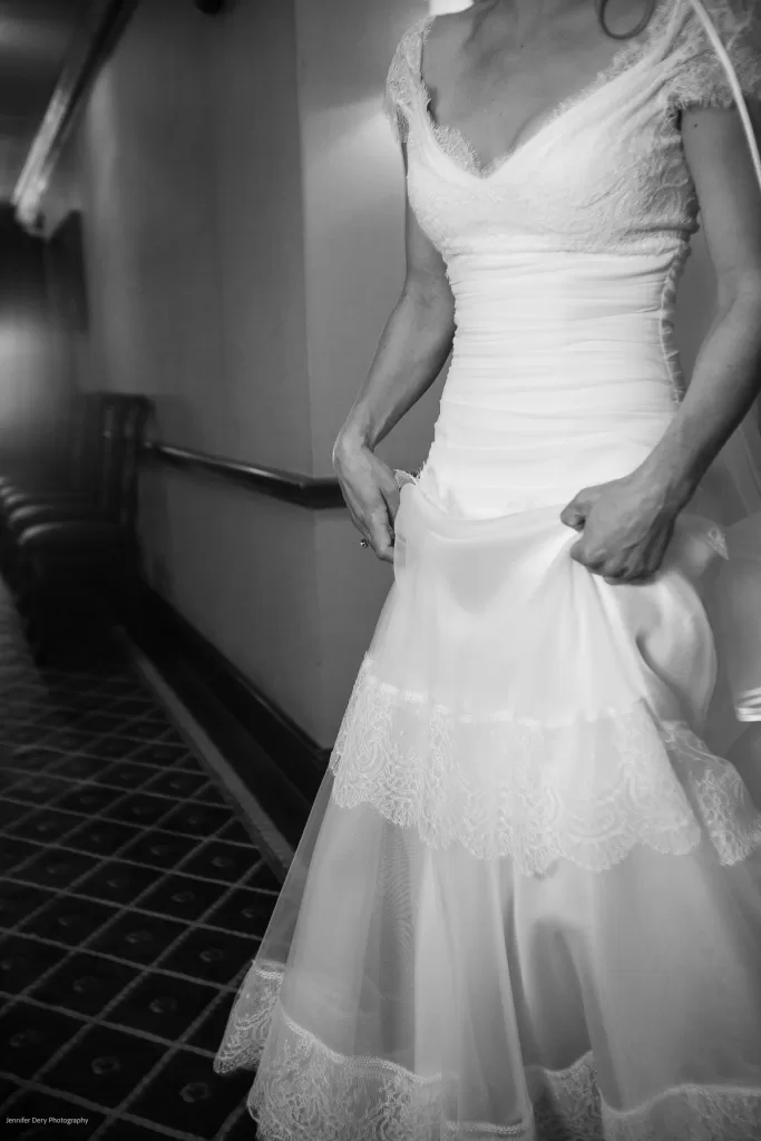 Black and white close-up of a bride in a wedding dress walking down a corridor. The dress has a lace bodice, short sleeves, and a layered skirt with lace trim. The bride is holding up part of her skirt as she walks, revealing delicate layers of fabric.