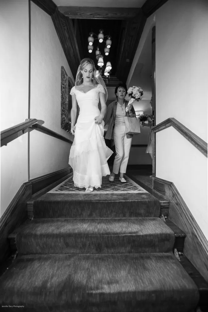 A woman in a wedding dress descends a staircase. Behind her, another person, dressed in a suit and holding a bouquet, follows. The setting seems to be indoors, with ornate woodwork along the stairwell. The image is in black and white.