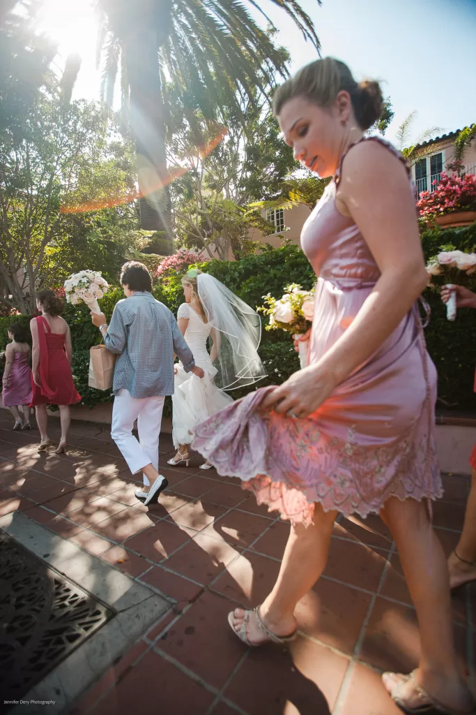 A bride and groom walk together while the bride, in a wedding gown and veil, shares a light moment looking at her groom. Bridesmaids in pink dresses surround them under the bright sun, walking on a tiled path with lush greenery and palm trees in the background.