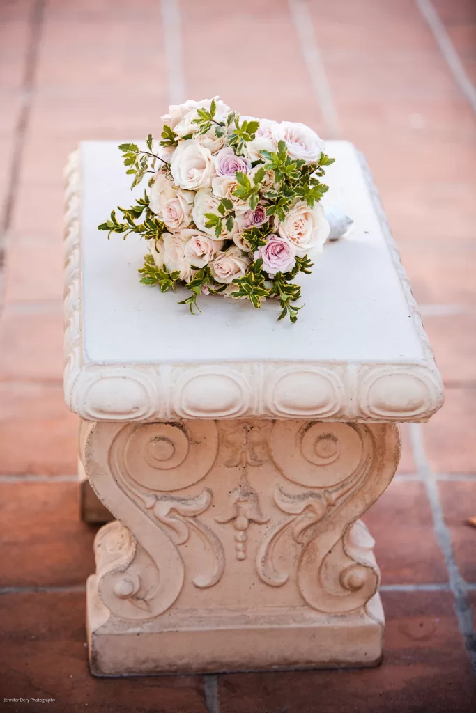 A bouquet of pink and white roses adorned with greenery rests on an ornate, cream-colored stone pedestal with intricate carvings. The pedestal sits on a terracotta tiled floor.