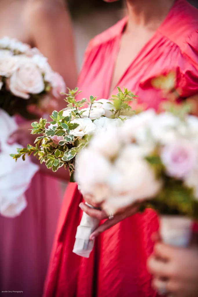 Close-up of two people holding bouquets of flowers. The focus is on the bouquets, which consist of white and pale pink roses with green foliage. One person is wearing a bright pink dress, while the other is wearing a lighter pink dress.