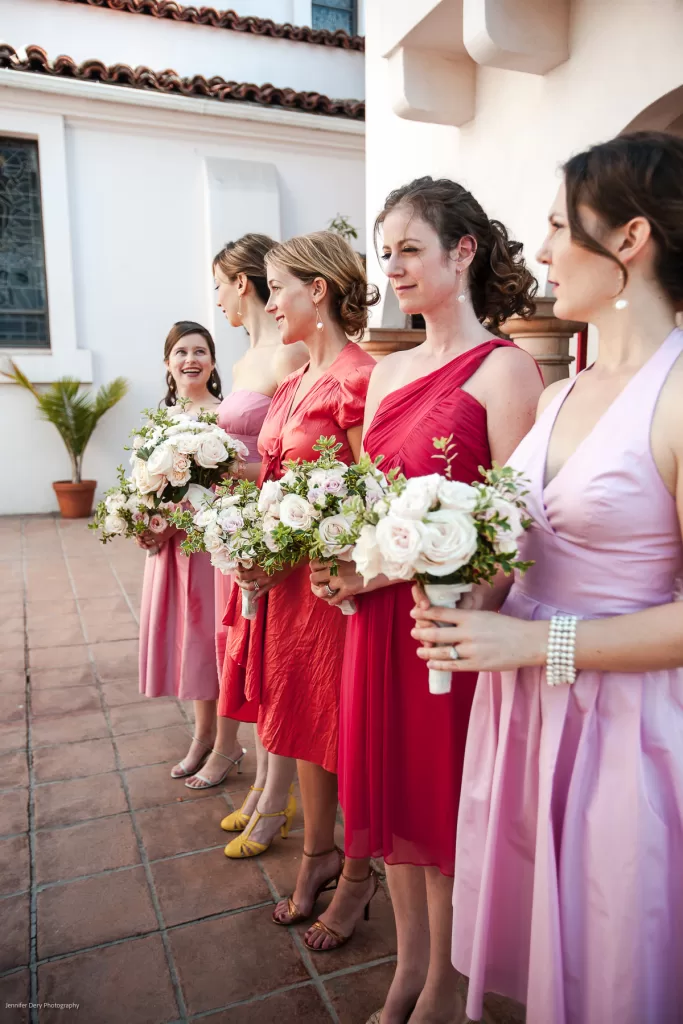 Five women dressed in various shades of pink and red bridesmaid dresses stand in a line outdoors. They each hold bouquets of white and pink flowers. One woman on the left looks at the others, smiling, while the rest look straight ahead.