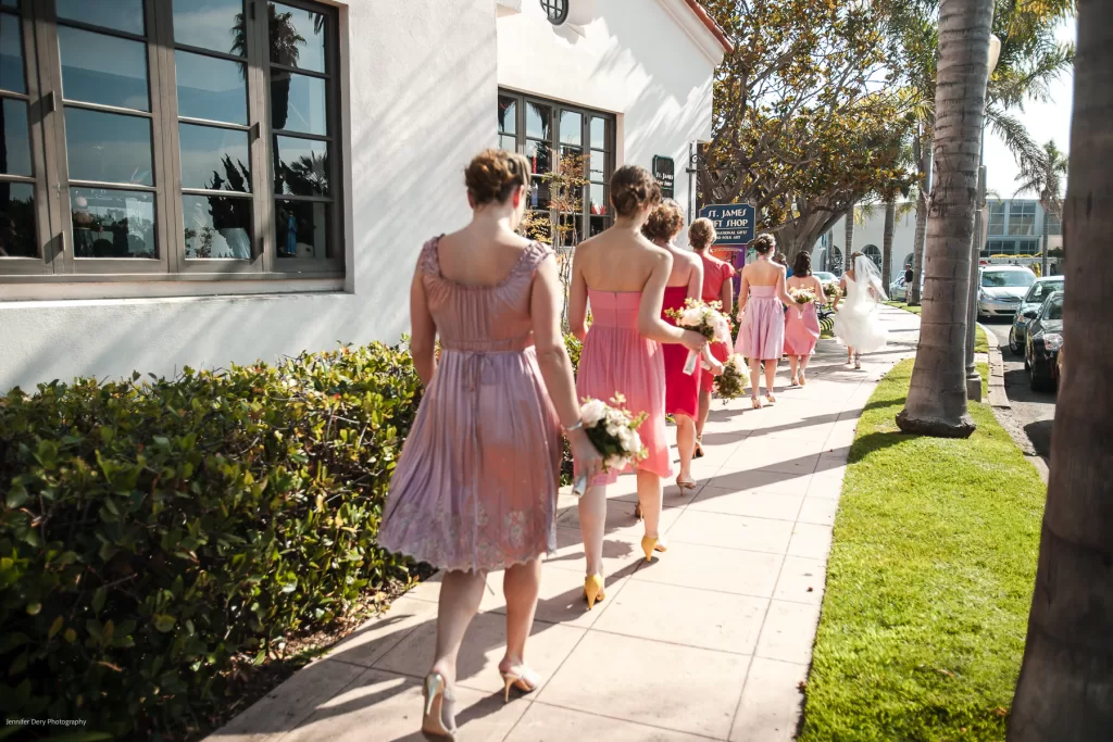 A group of women in pastel-colored dresses walk along a sunlit sidewalk next to a white building with large windows. Each woman is holding a bouquet of flowers. The scene appears festive, suggesting they might be bridesmaids heading to a wedding.