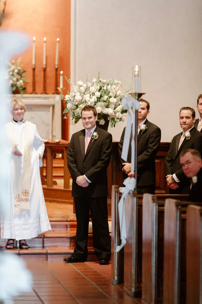 A groom, in a black suit and pink tie, stands at the altar beside three groomsmen. A woman in clerical attire stands next to them. A large floral arrangement is in the background. Guests are seated in pews, and white ribbons decorate the aisle.