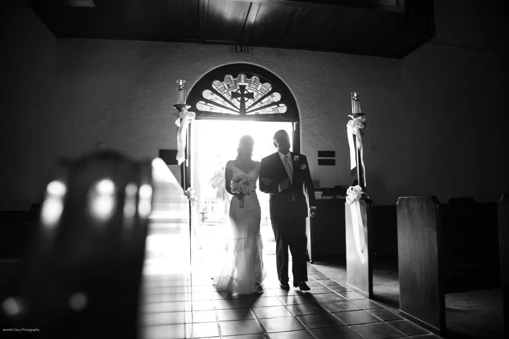 A bride holding a bouquet walks down the aisle with a man in a suit inside a dimly lit church. The bright sunlight streams through the open doors behind them, casting their silhouettes prominently. White ribbon decorations adorn the pews flanking the aisle.