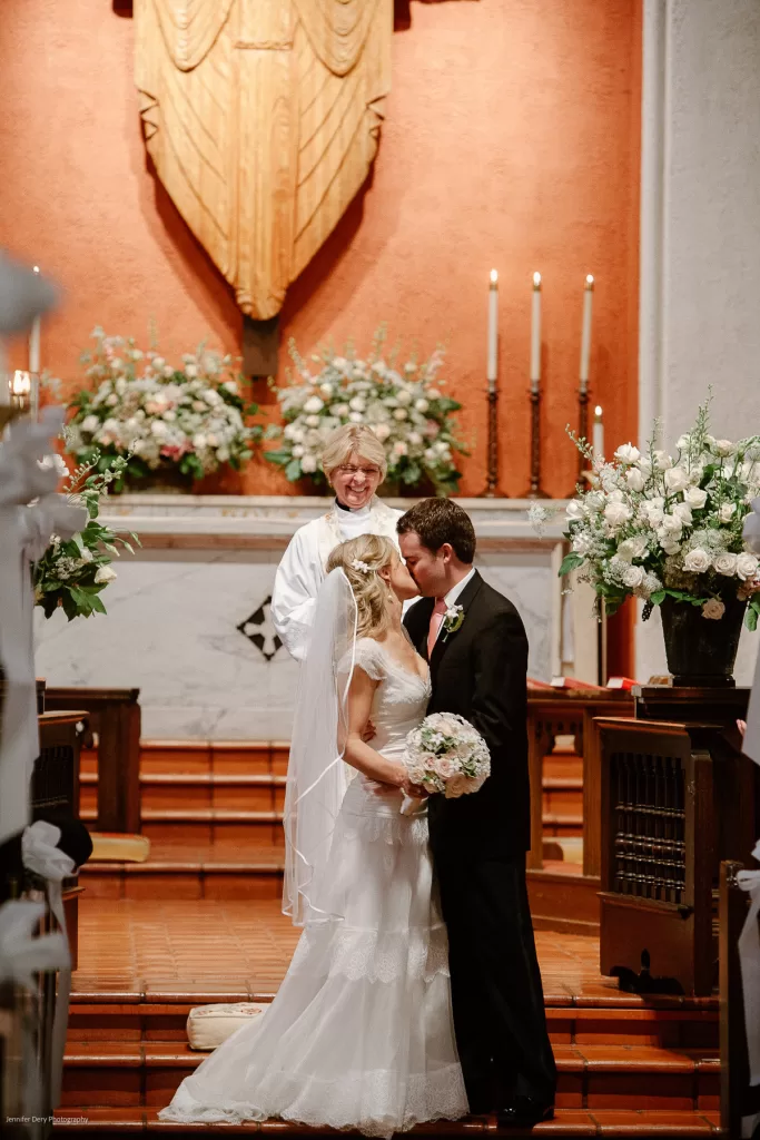 A bride and groom share a kiss at the altar while the officiant smiles behind them. The couple is wearing traditional wedding attire and the altar is adorned with flowers and candles.