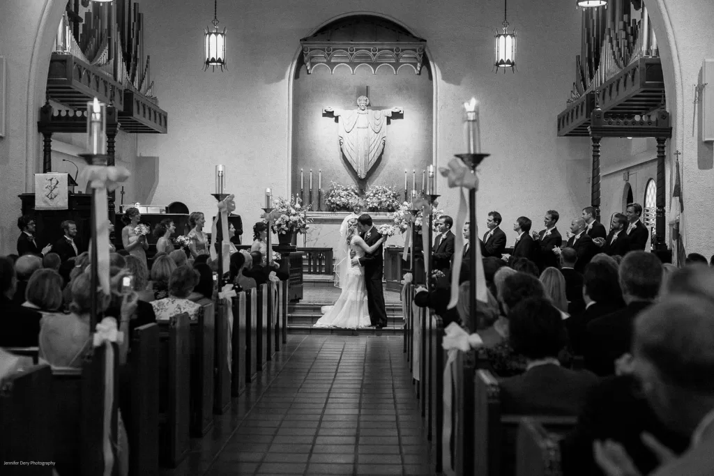 A black and white image of a wedding ceremony inside a church. The bride and groom share a kiss at the altar, surrounded by wedding attendants and guests seated in rows. The church features high ceilings, candlelit accents, and a statue behind the altar.