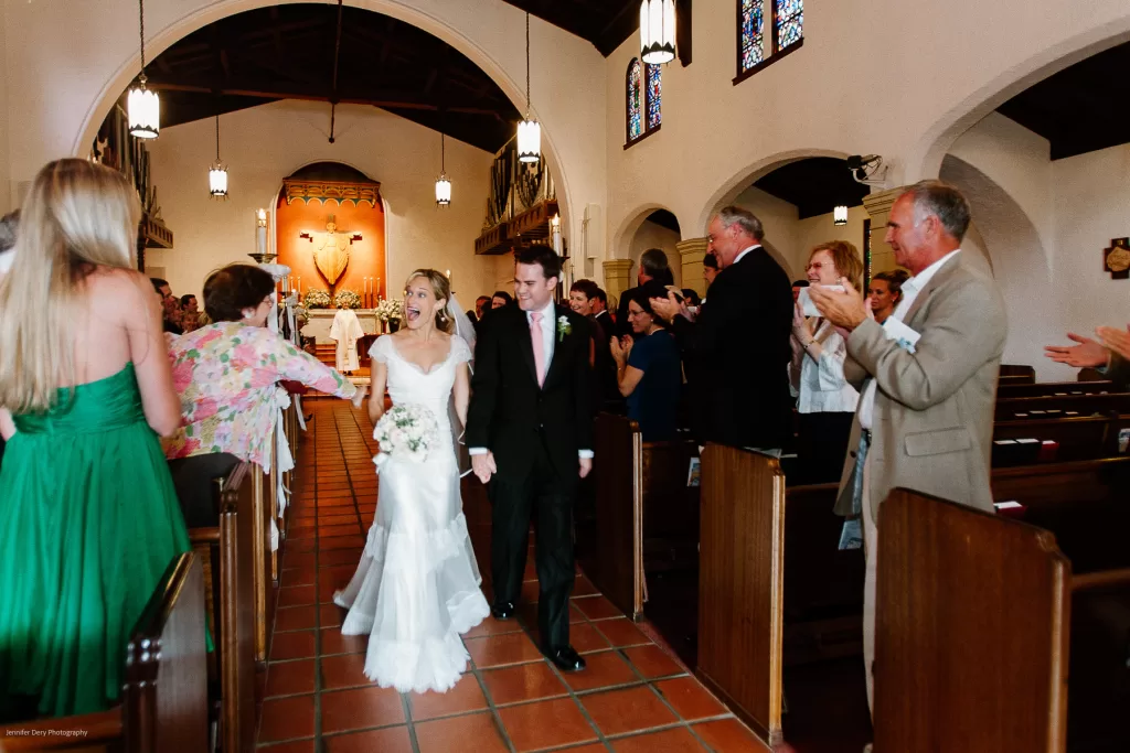 A bride and groom walk down the aisle of a church as guests applaud and smile. The bride wears a white gown and carries a bouquet, while the groom is in a black suit. The church has wooden pews, stained glass windows, and decorative lights.