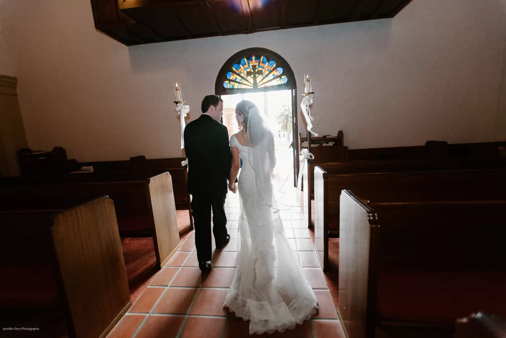 A bride and groom, dressed in a wedding gown and suit, walk hand-in-hand down the aisle of a church towards the bright light streaming through an ornate stained glass window above the entrance. The church pews are empty, adorned with small white floral arrangements.