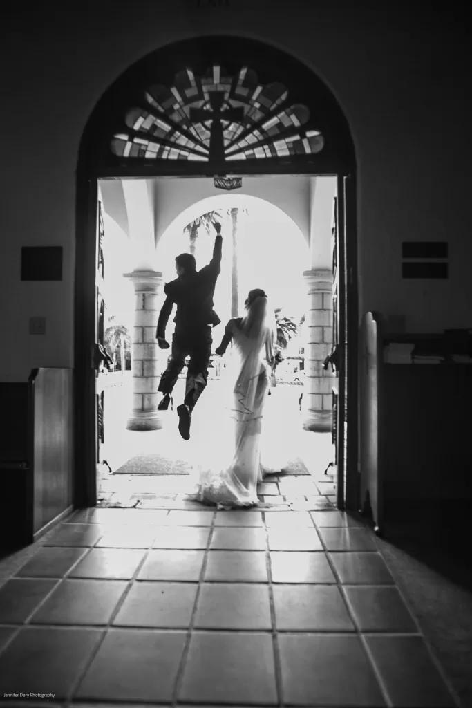 A bride and groom are holding hands, facing outside the entrance of a building with a tiled floor and arched doorway. The groom is captured mid-jump with one arm raised, while the bride stands beside him in a flowing gown.