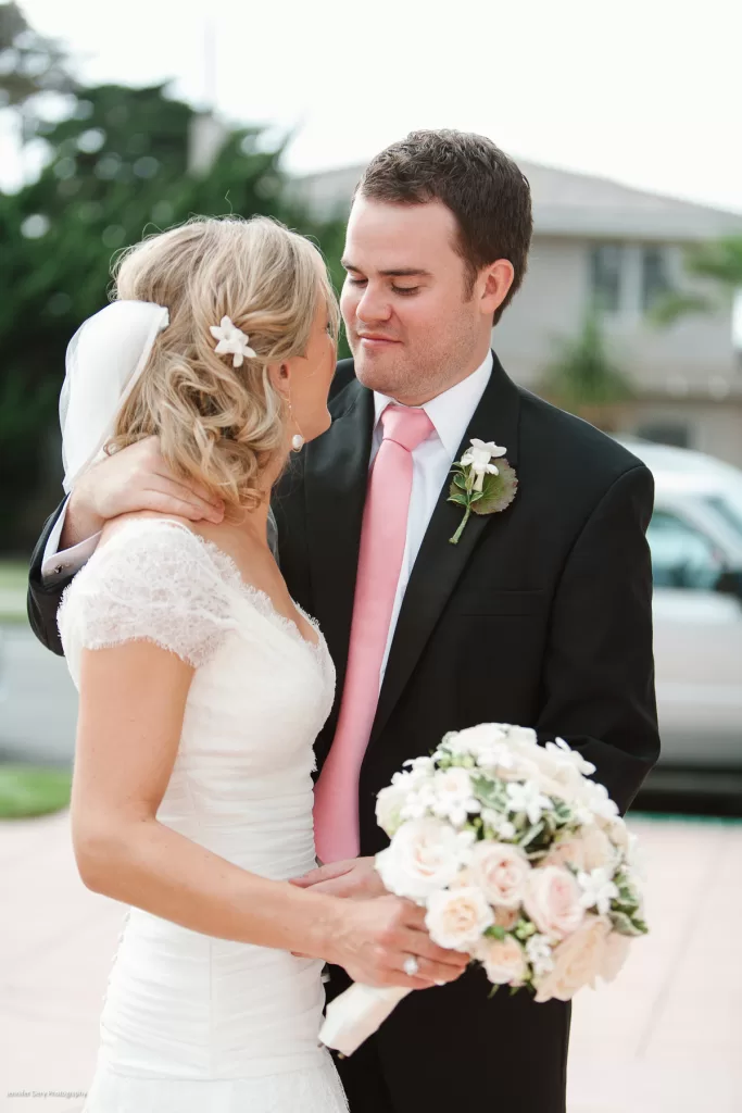 A bride and groom share an intimate moment on their wedding day. The bride, in a white lace dress and holding a bouquet of flowers, looks lovingly at the groom, who is wearing a dark suit with a pink tie. They are standing outdoors, with a house and car in the background.