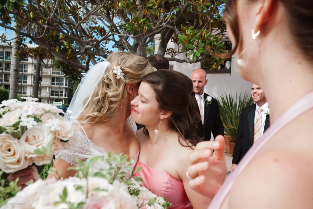 A bride in a white dress and veil shares a warm embrace with a woman in a pink dress, surrounded by lush greenery and buildings in the background. Another woman in pink is partially visible in the foreground, holding a bouquet of white flowers.