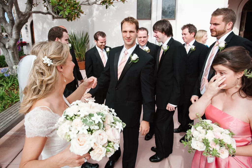 A bride in a white dress holding a bouquet of white flowers interacts with several groomsmen and a bridesmaid. The groomsmen are dressed in black suits with pink ties, and the bridesmaid wears a pink dress. They are standing outdoors.