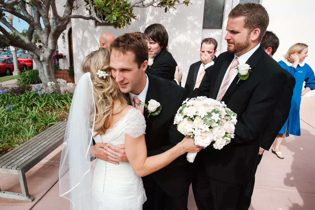 A groom and bride in wedding attire embrace under a tree. The groom holds a bouquet of white flowers. Several men in suits and boutonnieres stand nearby, one of them with crossed arms. In the background, a woman in a blue dress walks by.