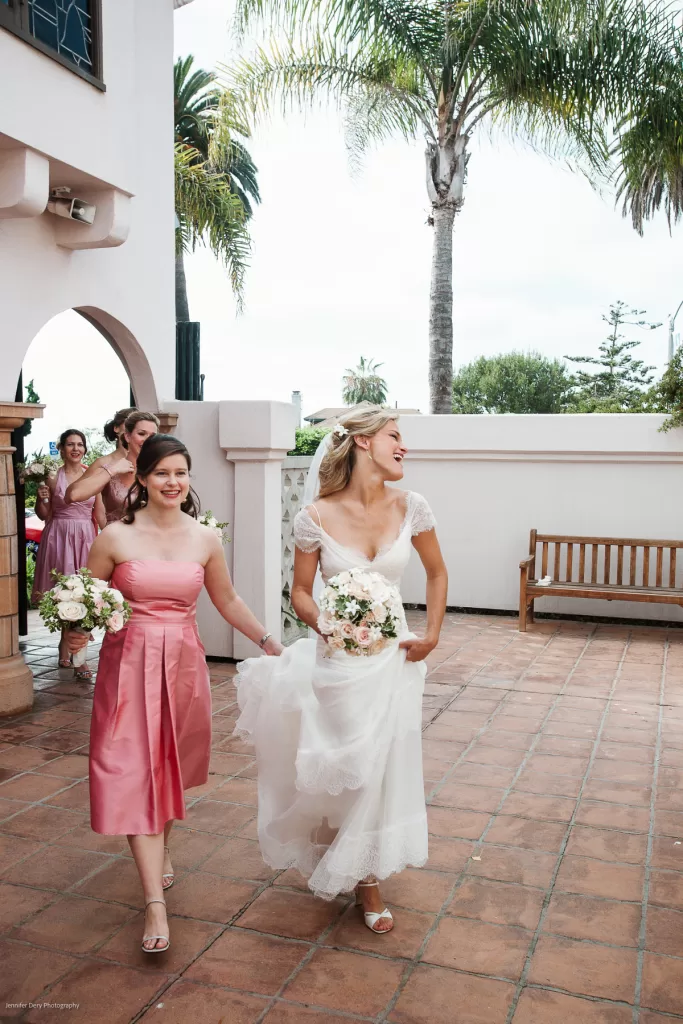 A bride in a white wedding dress and a bridesmaid in a pink dress walk joyfully outdoors. The bride holds her bouquet and part of her dress while the bridesmaid holds her own bouquet. Other bridesmaids in matching pink dresses are walking in the background. Lush greenery and a bench are visible.