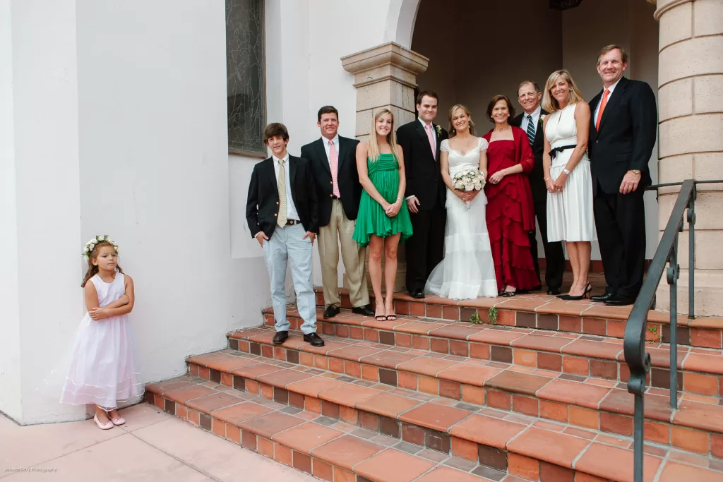 A group of people dressed in formal attire pose on the steps of a building, with a bride in a white dress holding flowers beside a groom in a black suit. An onlooker, a young girl in a white dress with a flower crown, stands to the left, separate from the group.