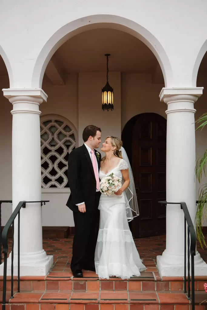 A couple stands together under an archway. The bride, in a white wedding dress holding a bouquet, gazes up at the groom, who wears a black suit with a pink tie. They are on a set of red brick steps in front of a white building with columns.